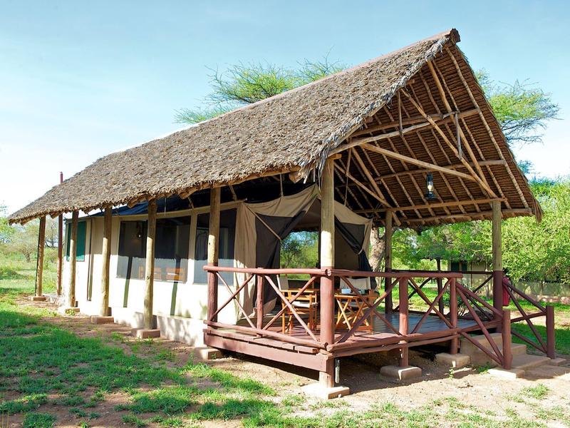 A simple safari tent with a thatched roof and porch in a green landscape.