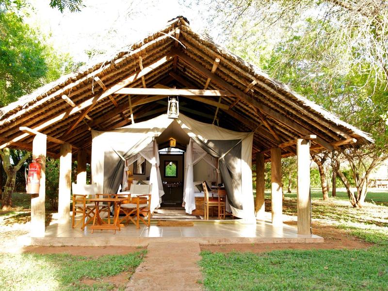 Open safari tent with wooden frame, chairs, and tables surrounded by trees and grass.