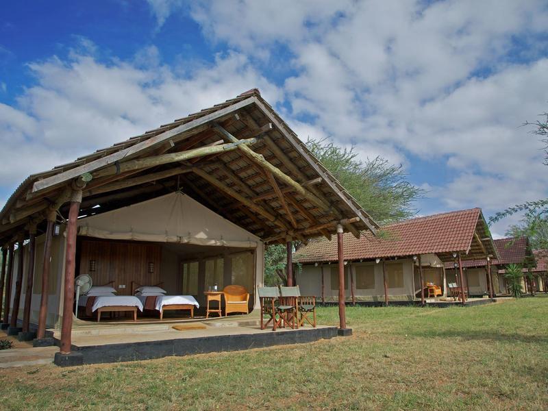 Single open lodge with beds and chairs surrounded by grass and trees under a blue sky.