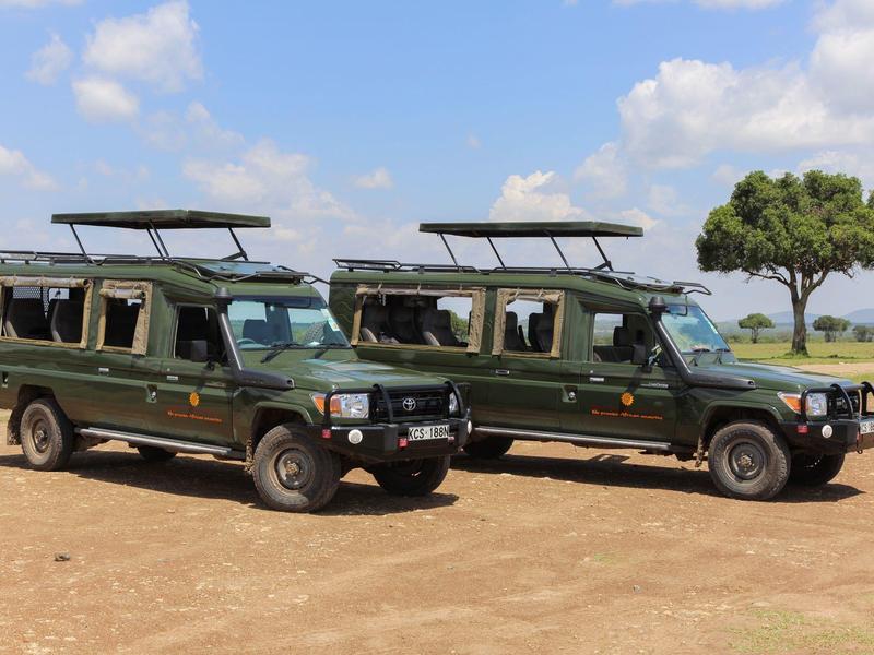 Zwei grüne Safari-Jeep-Fahrzeuge stehen auf trockenem Land unter blauem Himmel mit vereinzelten Wolken.
