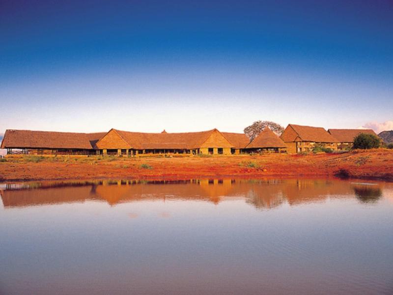 Edificio largo de hotel con techos rojos reflejado en agua tranquila bajo un cielo azul.