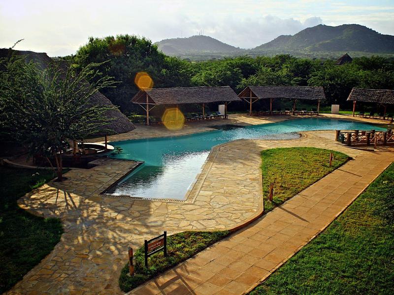 Zona de piscina natural con caminos, rodeada de montañas y pequeñas cabañas en el hotel.