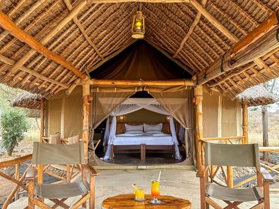 Geräumiges Safari-Zelt mit Holzstuhl, Bett und Getränken auf Veranda in Naturumgebung.