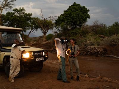 Zwei Männer mit Kamera neben einem Safari-Jeep in einer trockenen, bewaldeten Landschaft.