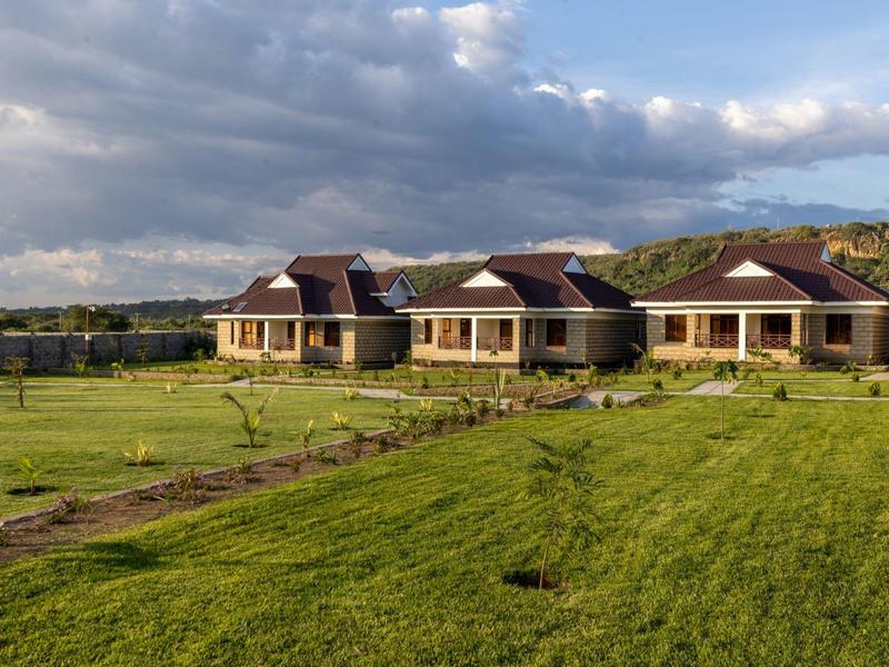 Three small houses with brown roofs on a vast green field under a cloudy sky.