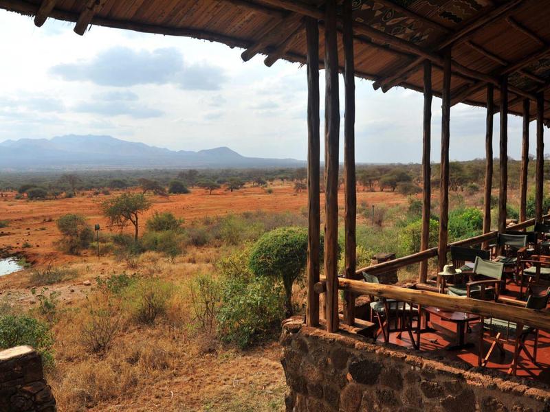 Open veranda with wooden tables and chairs overlooking wide, dry savannah and mountains.