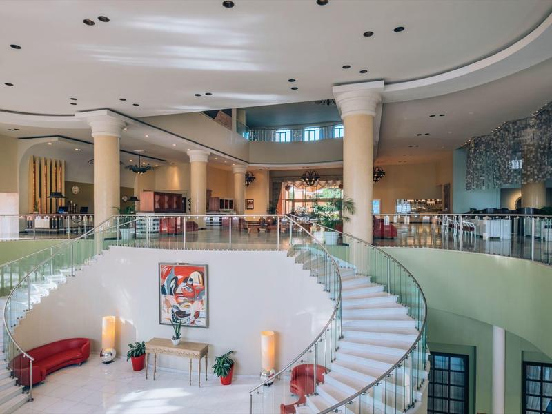 Vestíbulo de hotel luminoso con escalera curva, columnas y modernos asientos en rojo y blanco.