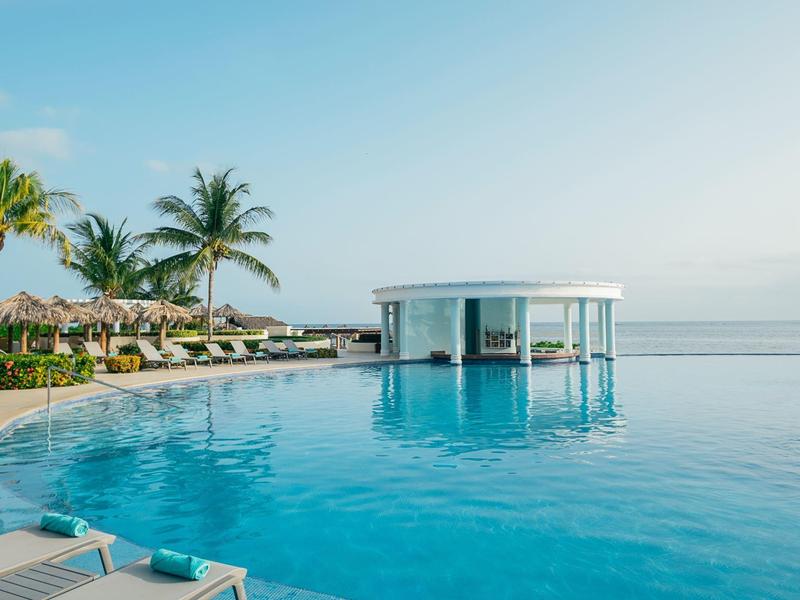 Piscina infinita moderna con bar, rodeada de palmeras y tumbonas en un día soleado.