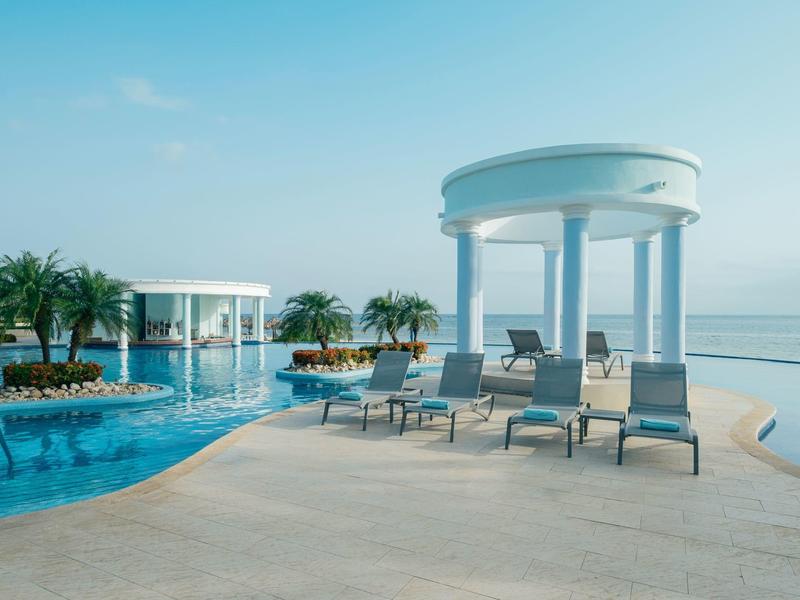 Piscina infinita con tumbonas y pabellón blanco junto al mar tranquilo bajo cielo azul.