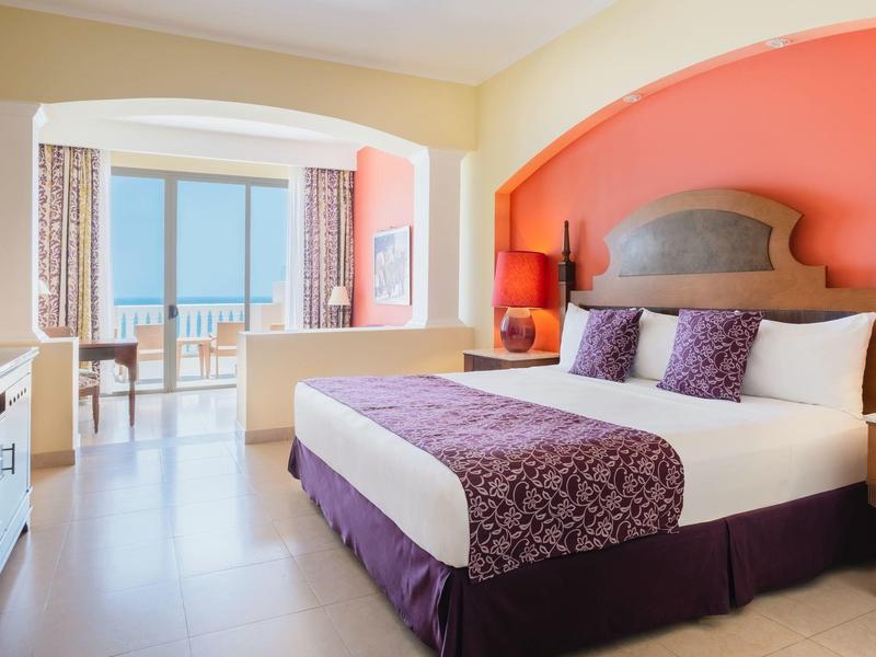 Habitación de hotel luminosa con cama grande, balcón y vista al mar al fondo.