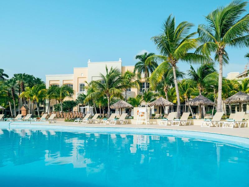Large pool with sun loungers and palm trees in front of hotel buildings under clear sky.