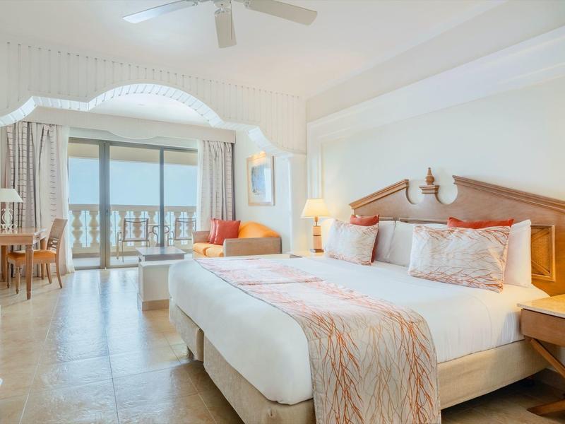 Bright hotel room with large bed, seating area, and balcony overlooking the sea.