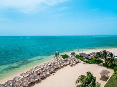 Strand mit weißen Sonnenschirmen und klarblauem Wasser neben tropischem Hotelgebiet.