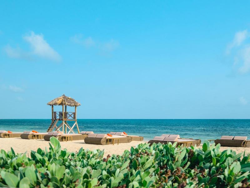 Pláž s zelenými keři, dřevěnými lehátky a malým altánem pod jasnou oblohou.