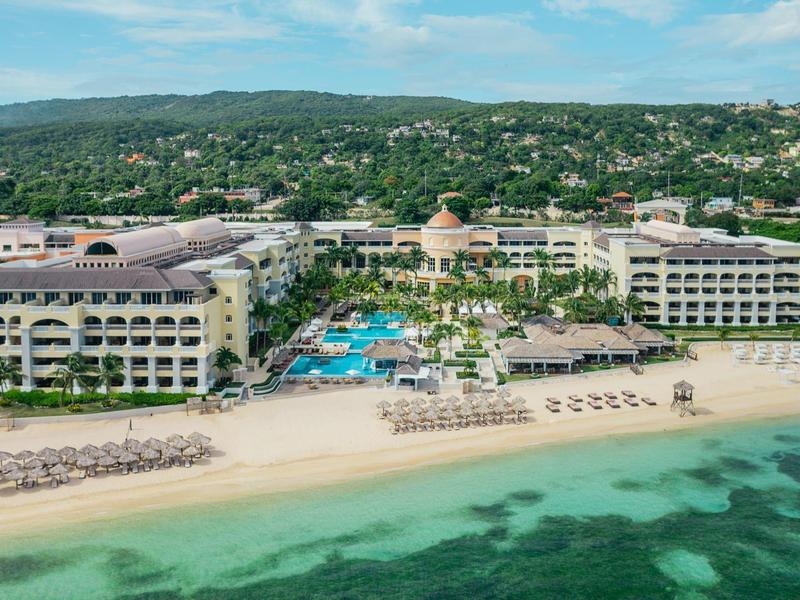 Luxusní plážový resort s bazénem, lehátky a zelenými kopci v pozadí.