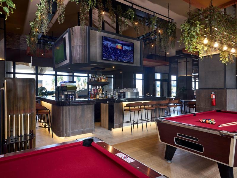 Moderne Bar mit Holzmöbeln, grünen Pflanzen und roten Billardtischen im gemütlichen Ambiente.