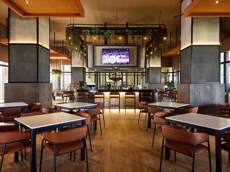 Modernes Restaurant mit Holzboden, vielen Tischen, braunen Stühlen und einem beleuchteten Barbereich.