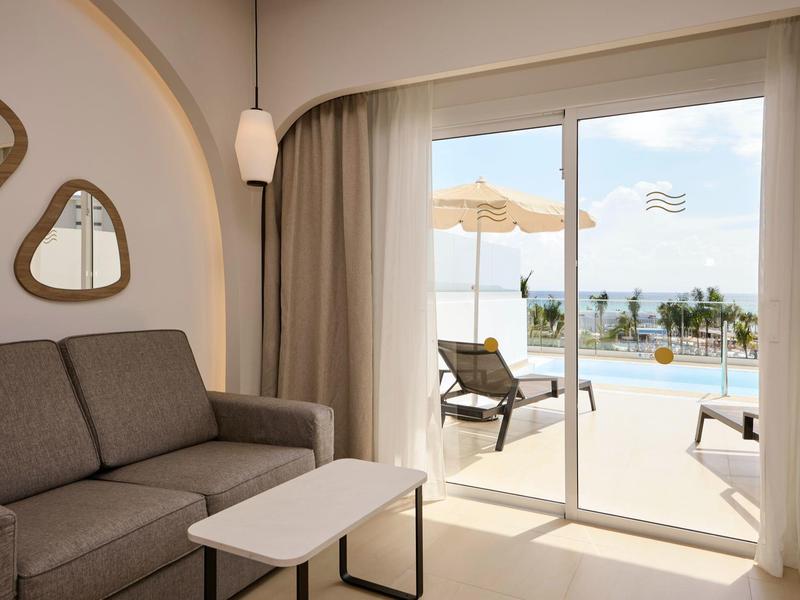 Helles Wohnzimmer mit grauem Sofa, weißem Tisch und Blick auf Terrasse mit Pool und Meer.
