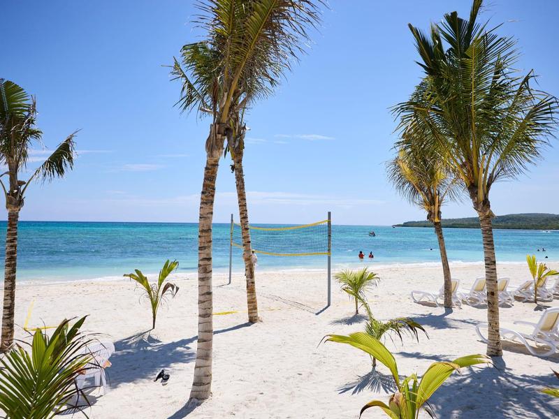 Strand mit weißem Sand, Palmen und klarem, blauem Meer unter hellem Himmel.