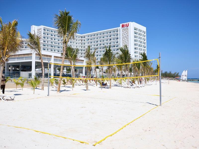 Weißer Sandstrand mit Volleyballnetz, Palmen und großem Hotelgebäude im Hintergrund bei klarem Himmel.