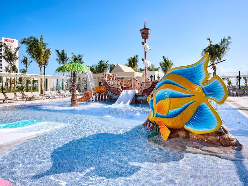 Bunter Kinderspielplatz mit Wasser, großer Fischrutsche und Palmen unter blauem Himmel.