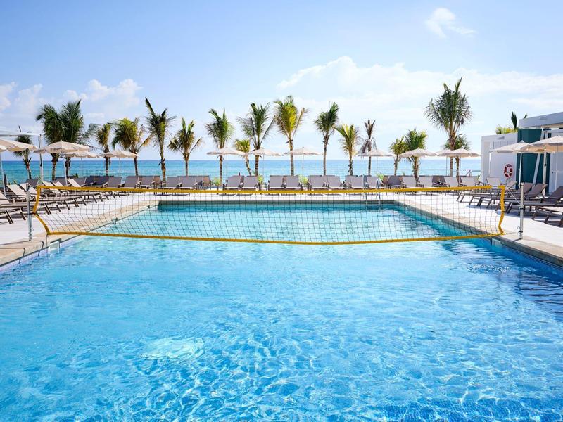 Großer, klarer Pool mit Liegestühlen und Palmen am Rand, blauer Himmel im Hintergrund.