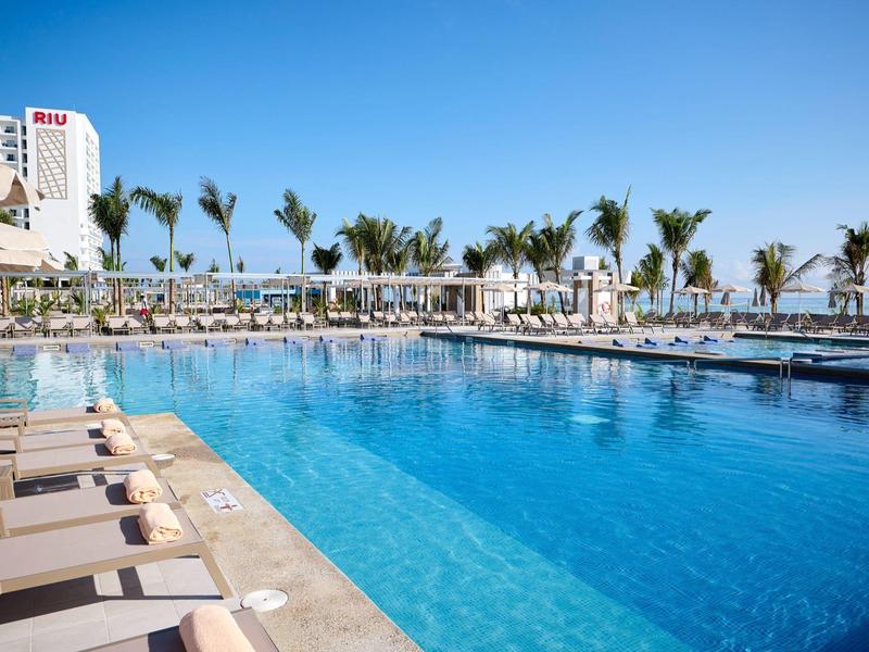 Großer Pool mit klar blauem Wasser, Liegen am Rand und Palmen im Hintergrund unter klarem Himmel.