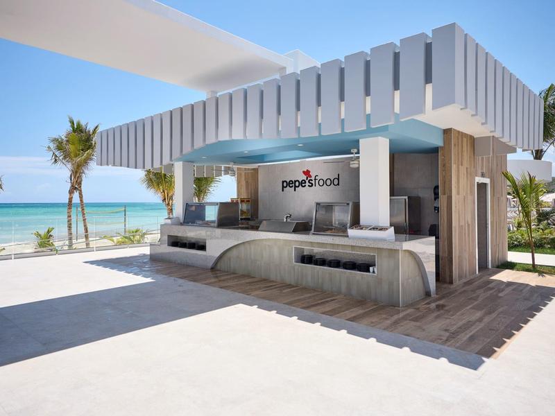 Modernes, offenes Strandrestaurant mit Beton, Holz, Palmen und Meer im Hintergrund unter blauem Himmel.