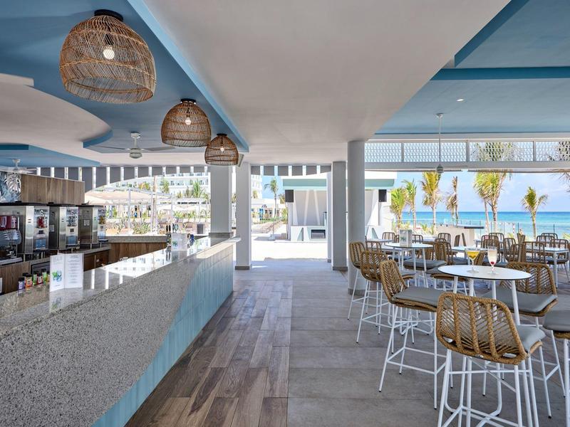 Moderner Cafébereich mit Bar, Holzstühlen, großen Fenstern und Blick auf die Uferpromenade.