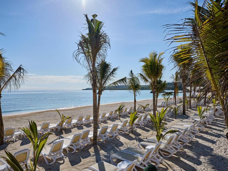 Strand mit weißen Liegestühlen, Palmen und ruhigem blauem Meer unter klarem Himmel.