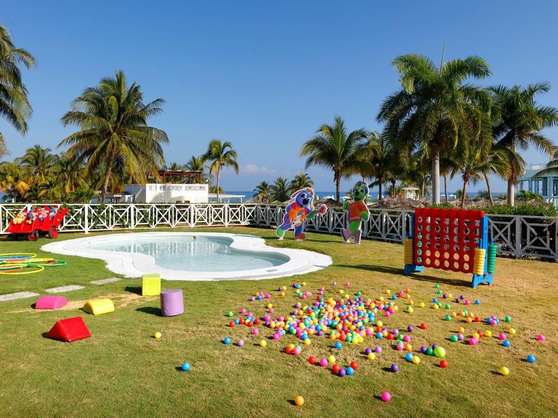 Zona de juegos al aire libre con piscina pequeña, césped y árboles tropicales bajo cielo despejado