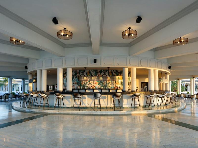 Bar moderna de hotel con barra circular, lámparas colgantes y taburetes en ambiente claro.