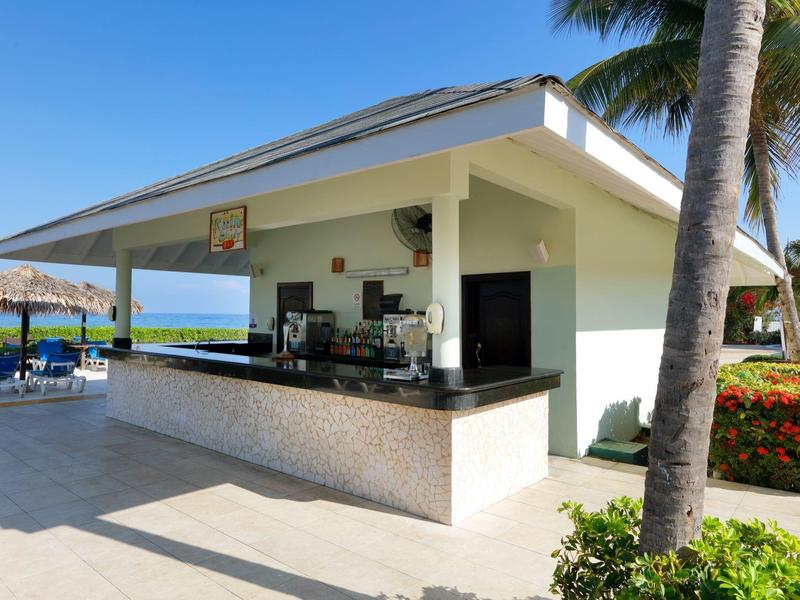 Área moderna de bar junto a la piscina con vista al mar, palmeras y sombrillas bajo cielo despejado.