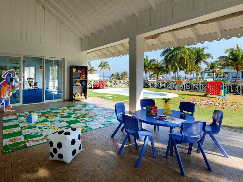 Kinderspielecke mit blauem Tisch, bunten Stühlen, Spielfeldteppich, Würfelhocker und Palmen im Hintergrund.