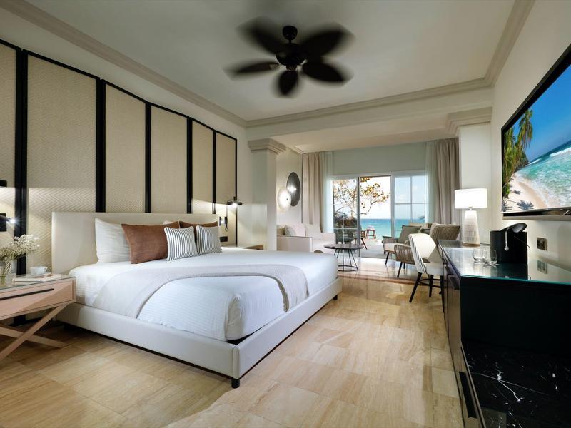 Helles Hotelzimmer mit großem Doppelbett, Deckenventilator, Schreibtisch, Fernseher und Meerblick.