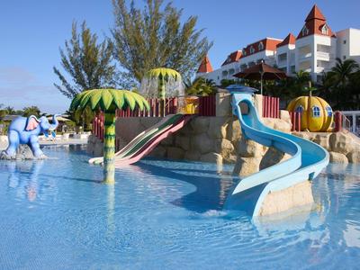 Bunter Kinderspielplatz mit Rutschen und Wasserspielen in flachem Pool vor einem Hotel.