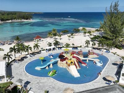 Großer Pool mit bunten Wasserrutschen, umgeben von Liegestühlen, Palmen und Meer im Hintergrund.