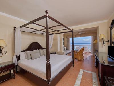 Großes Zimmer mit Himmelbett, zwei Sesseln, TV, warmen Farben und Meerblick vom Balkon.