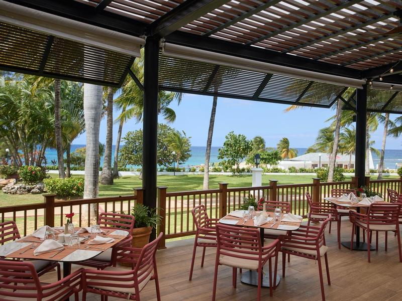 Zastřešená terasa s stoly a židlemi s výhledem na palmy a moře v hotelovém resortu.