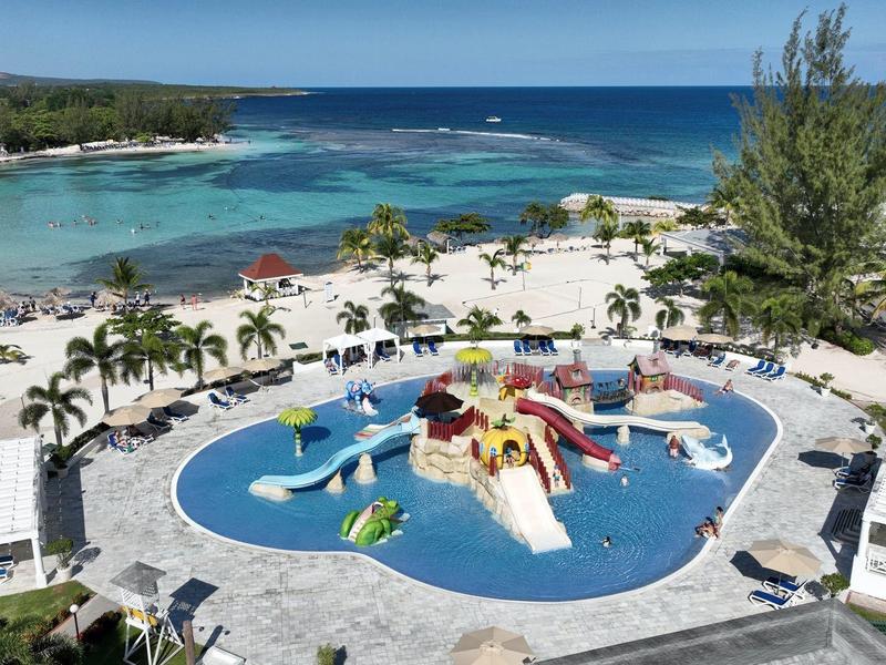 Bazén s vodní skluzavkou v tropickém letovisku u moře s palmami a lehátky.