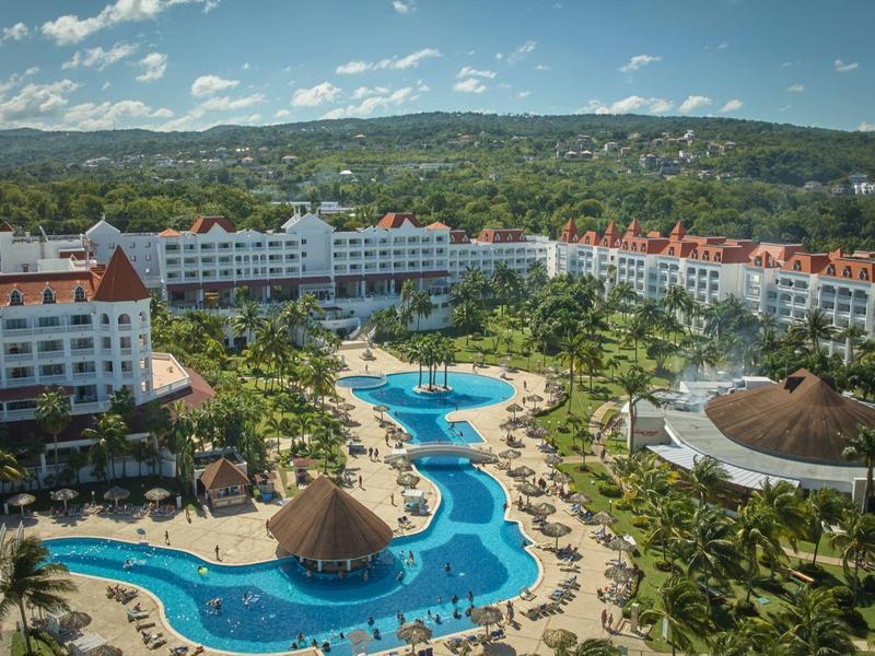 Velké letovisko s několika bazény, palmami a vícepodlažními hotelovými budovami u zelených kopců.