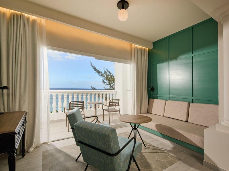 Světle zařízený hotelový pokoj s posezením a balkonem s výhledem na moře.