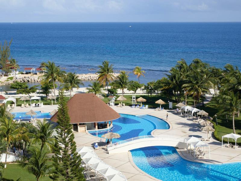 Hotelový bazén s palmami a výhledem na oceán pod modrou oblohou.