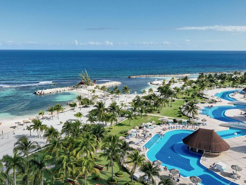 Letecký pohled na tropické pobřeží s palmami, bílou písečnou pláží a klikatým bazénem.