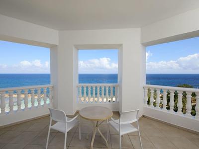 Balkon mit weißen Stühlen und Tisch, mit Meerblick unter blauem Himmel und einigen Wolken.