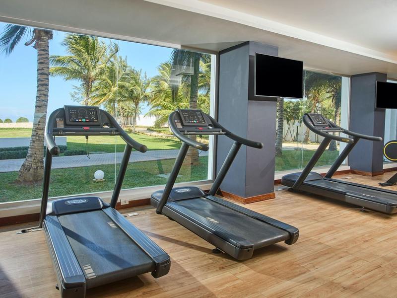 Fitnessruimte met loopbanden en uitzicht op palmbomen en zee door grote ramen.