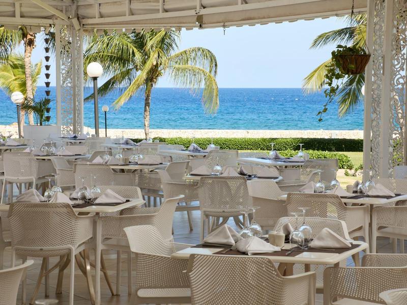 Open restaurant met witte stoelen en tafels met uitzicht op de oceaan en palmbomen.