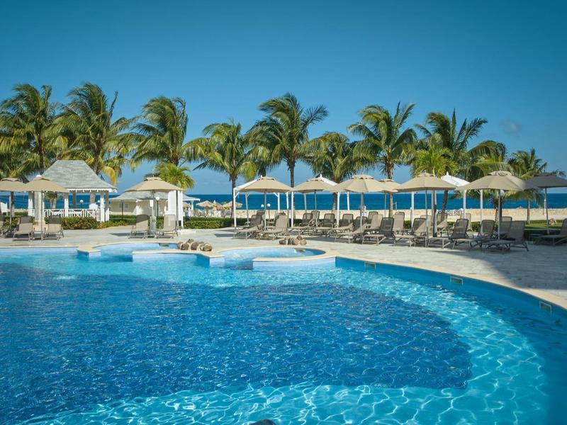 Groot zwembad met parasols en palmbomen onder een heldere hemel bij een strandhotel.