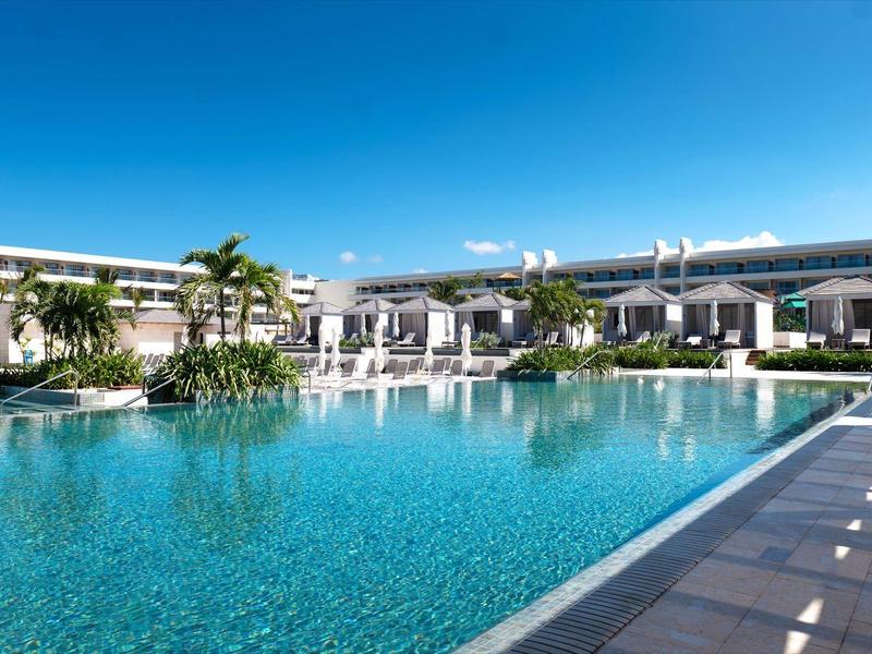 Großer Pool in modernem Hotel mit klar blauem Wasser, Sonnenschirmen und grünen Palmen.