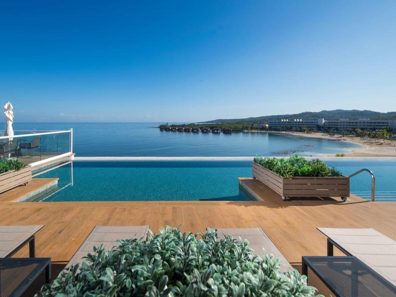Moderner Pool mit Holzdeck und Meerblick, blauer Himmel und Sandstrand im Hintergrund.